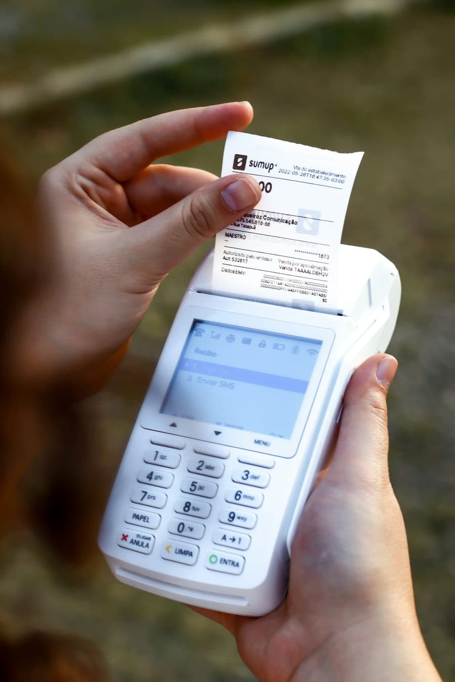 Mão segurando a maquininha de cartão com o recibo saindo, sem letra miúda